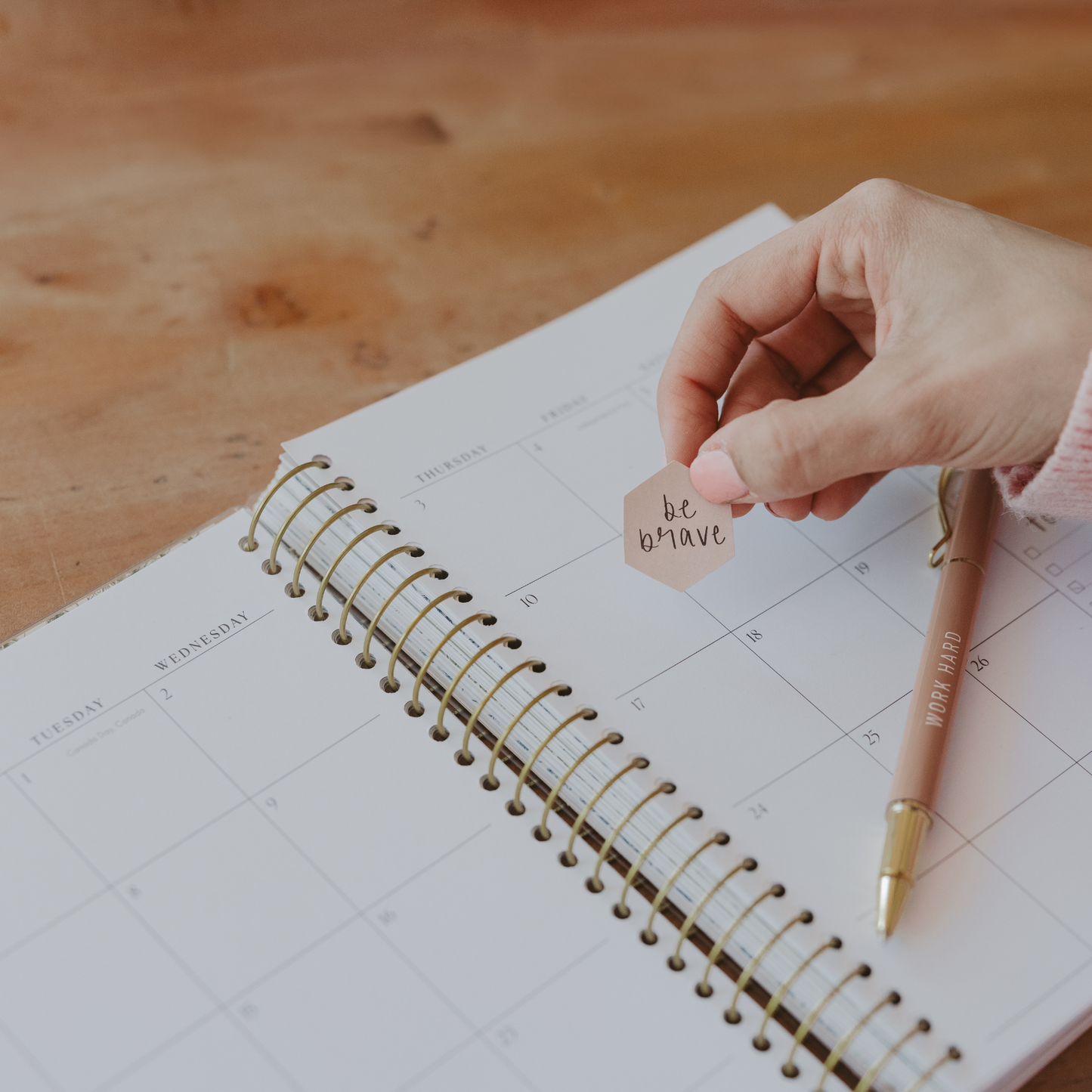 Person holding a 'be brave' tag over an open planner on a wooden surface