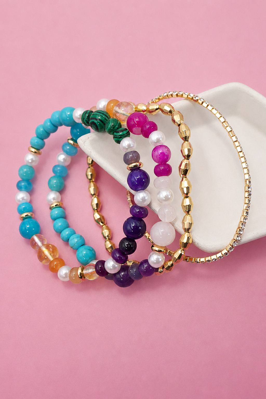A set of five colorful beaded bracelets with gold-tone and white-tone elements, arranged on a white ceramic ring stand against a solid pink background.