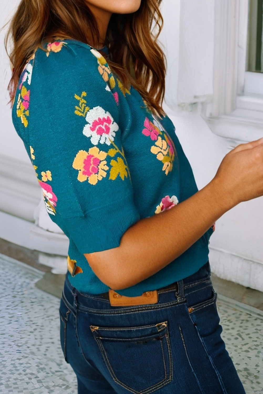 Person wearing a teal blouse with colorful floral patterns and blue jeans indoors.