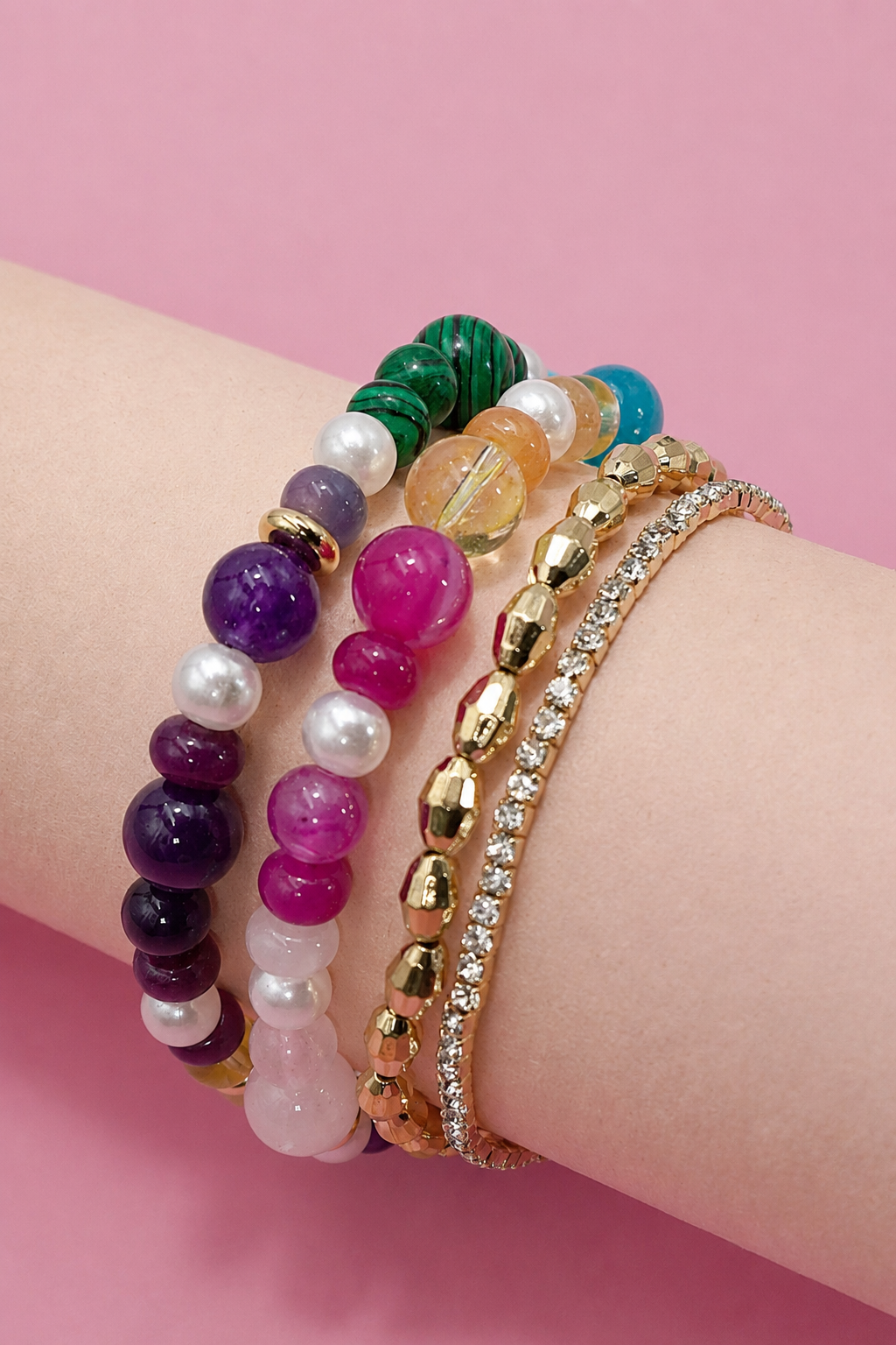 A set of five colorful beaded and stone bracelets stacked on a person's wrist, featuring purple, pink, green, white, and gold-toned pieces, set against a solid pink background.