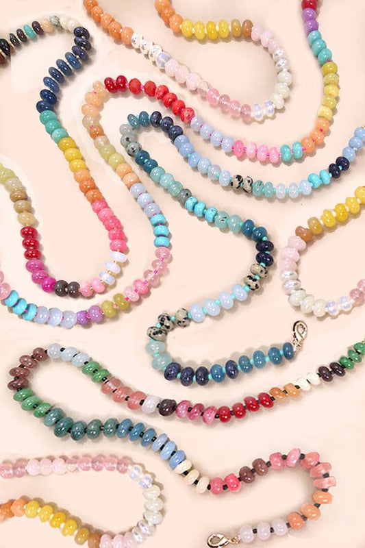 A collection of colorful beaded necklaces and bracelets arranged in loops on a light peach background. The beads come in many different colors including pink, blue, yellow, green, orange, and purple, with some pieces featuring iridescent finishes and small silver-toned metal spacers.